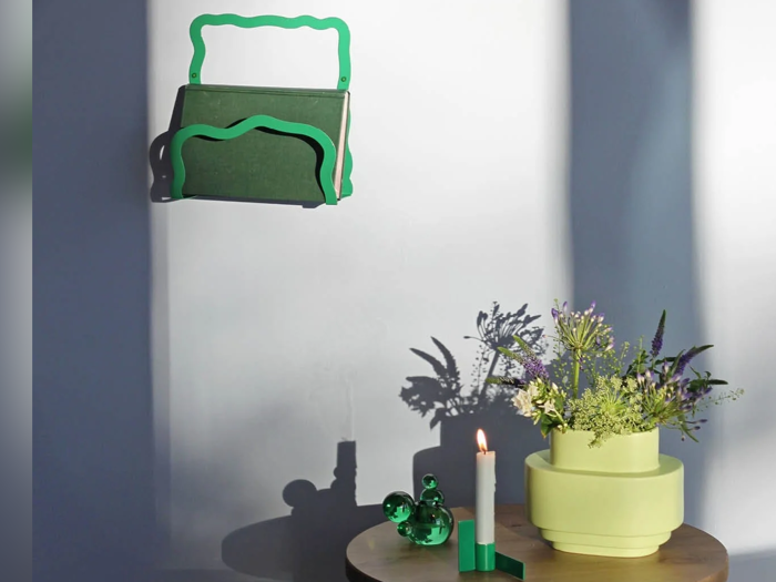 En grøn bogreol med bøger er monteret på en hvid væg over et træbord med en gul blomstervase, et tændt stearinlys og grøn glasdekoration, som kaster tydelige skygger.