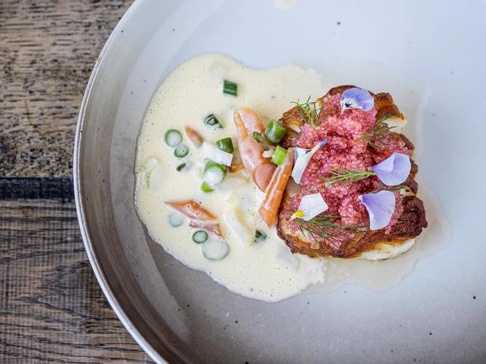 En tallerkenanrettet gourmetret med stegt fisk med rosa rogn, spiselige blomster og urter, serveret med cremet sauce, grøntsager i skiver og garniture på et rustikt træbord.