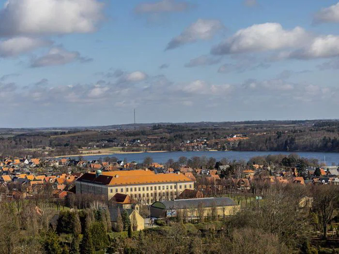 En by med orangefarvede bygninger, en sø og træer i forgrunden under en delvis overskyet blå himmel.