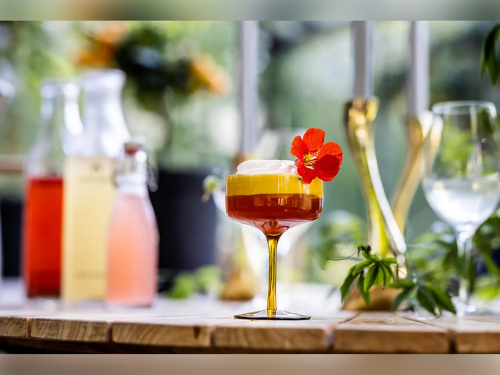 En farverig lagdelt cocktail i et glas pyntet med en rød spiselig blomst står på et træbord med flasker og planter sløret i baggrunden.