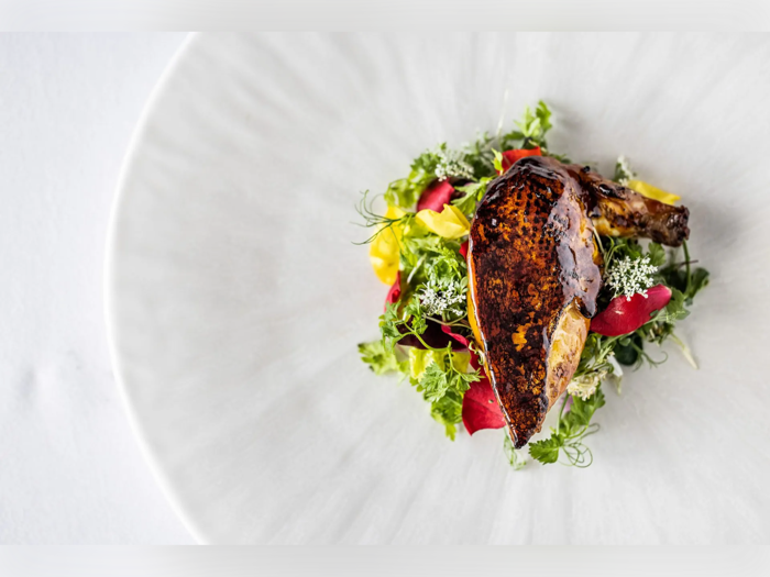 En gourmetret med glaseret stegt kød på en bund af frisk grønt, krydderurter og farverige spiselige blomster, præsenteret på en hvid tallerken.