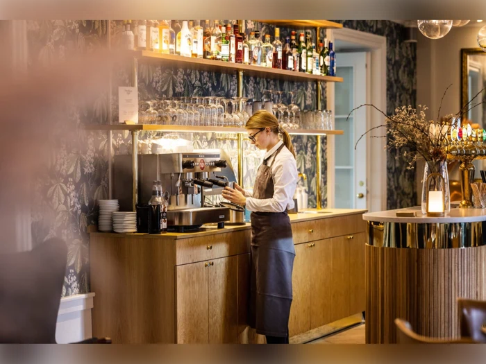 En barista i forklæde tilbereder en drink ved en kaffemaskine bag en trædisk med hylder med glas og flasker i en stilfuld café.