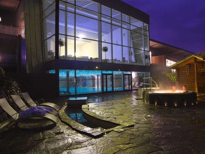 En moderne spa om natten med store glasvinduer, en indendørs pool, liggestole og to personer, der slapper af i et rundt udendørs boblebad.