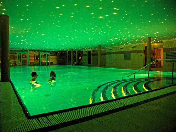 To personer svømmer i en stor indendørs pool med glødende grønt vand og et loft dekoreret med stjernelignende lys. Buede trin fører ned i poolen.