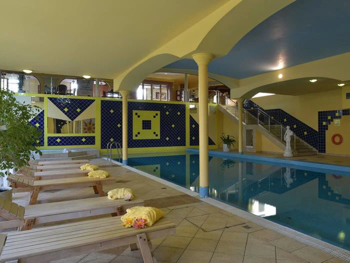 Indendørs poolområde med liggestole i træ, gule håndklæder, en statue, blå og gule flisevægge og en trappe, der fører op til et øvre niveau.
