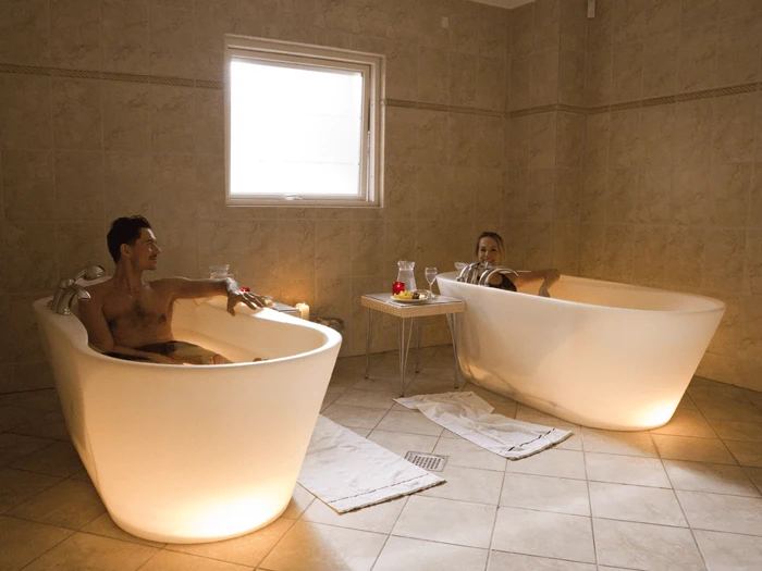 To personer slapper af i hvert sit badekar i et flisebadeværelse med håndklæder på gulvet, et bord med drikkevarer og tændte stearinlys ved vinduet.
