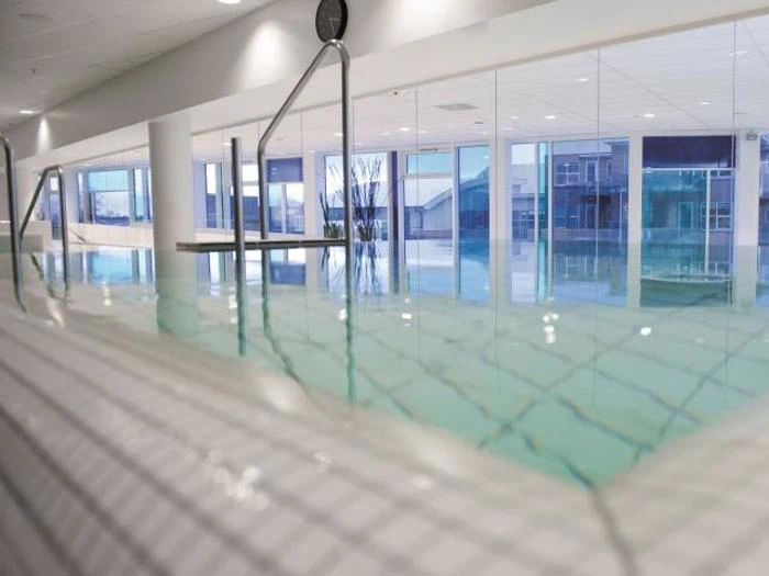 Indendørs swimmingpool med klart vand, flisebelagte kanter, metalgelændere og store vinduer, der lukker naturligt lys ind. Moderne, lyst og rent interiør.