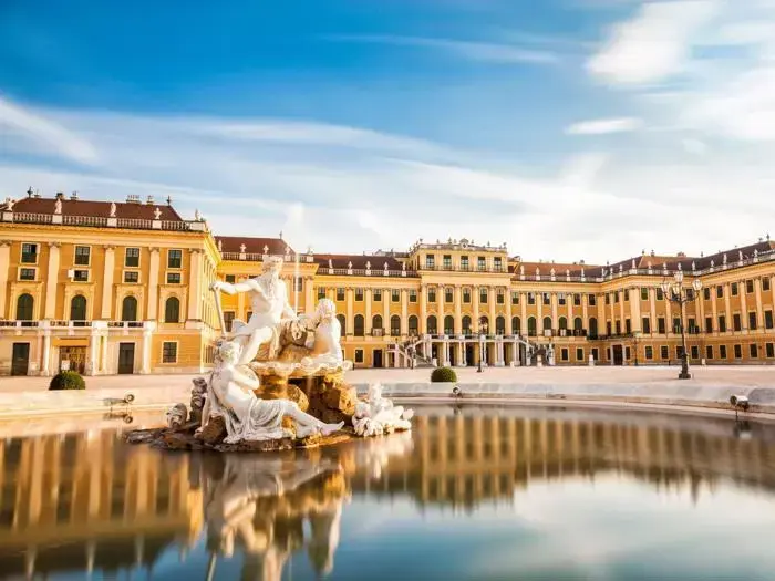 Barokpalads med gul facade og mange vinduer, bag et stort springvand med klassiske statuer og klart vand, under en blå himmel med tynde skyer.