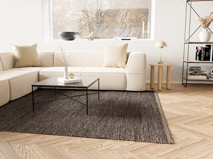 Minimalistisk stue med cremefarvet sektionssofa, sofabord i sort metal, gråt tæppe og lyst trægulv. Stort vindue med træer udenfor og hylder med indretning i baggrunden.