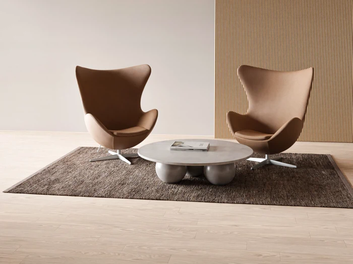 To moderne brune loungestole står over for hinanden på et rundt gråt sofabord på et brunt tæppe i et minimalistisk rum med lyse trægulve og en væg med træpaneler.
