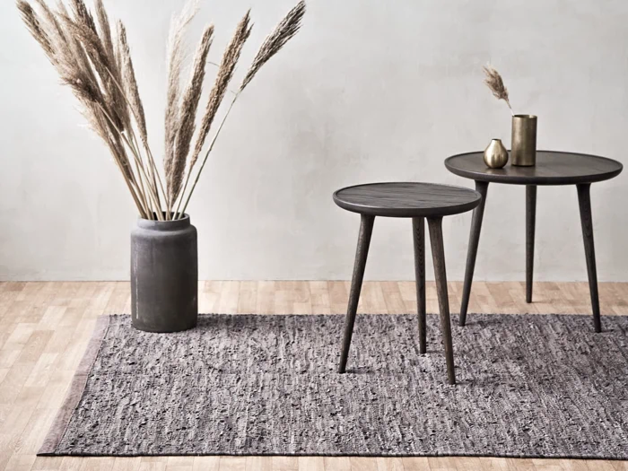 Minimalistisk indretning med to runde mørke træborde, et gråt tekstureret tæppe, en stor grå vase med højt tørret græs og pyntegenstande i messing på et lyst trægulv.