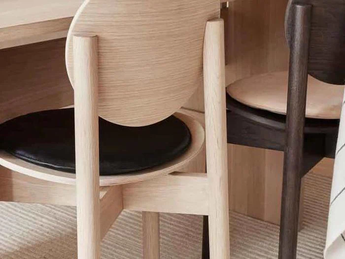 To træstole med runde rygge og sorte sædehynder er placeret ved siden af et træskrivebord på et lyst, struktureret tæppe.