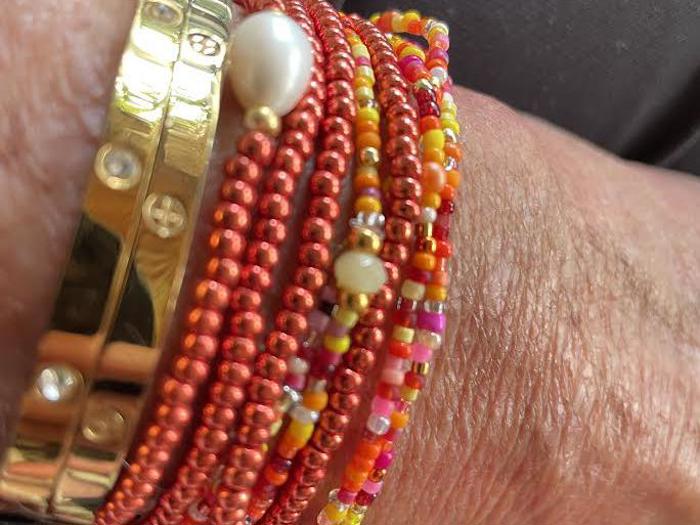 Nærbillede af et håndled med guldarmbånd og flere perlearmbånd i orange, rød, gul, pink og hvid, med en perle på et af perlearmbåndene.