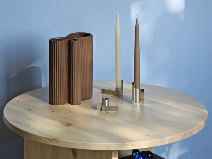 Et rundt træbord med en brun, bølget vase, to høje stearinlys i moderne stager og en lille cylindrisk genstand mod en lyseblå væg.