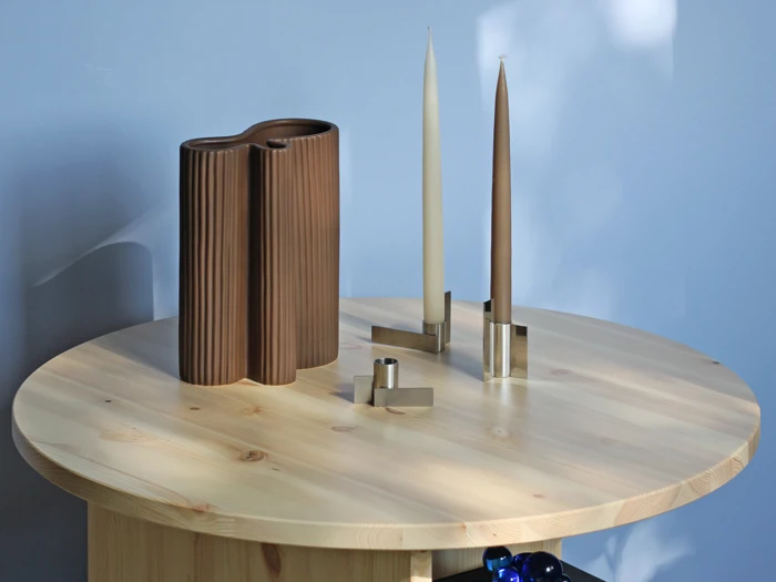 Et rundt træbord med en brun, bølget vase, to høje stearinlys i moderne stager og en lille cylindrisk genstand mod en lyseblå væg.