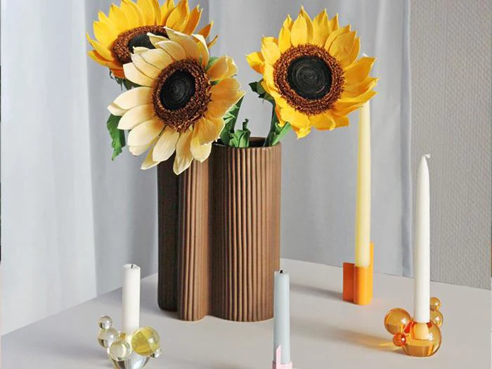 En brun, bølget vase rummer tre store solsikker, omgivet af fire farverige lys i moderne, geometriske stager på en hvid flade.