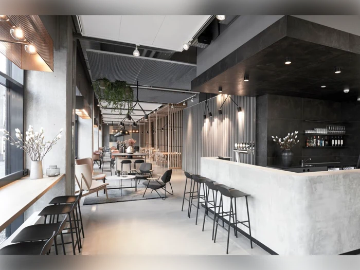 Moderne minimalistisk caféindretning med betonbar, sorte taburetter, loungestole, store vinduer og blød belysning. Dekorative planter og neutrale toner skaber en elegant og indbydende atmosfære.