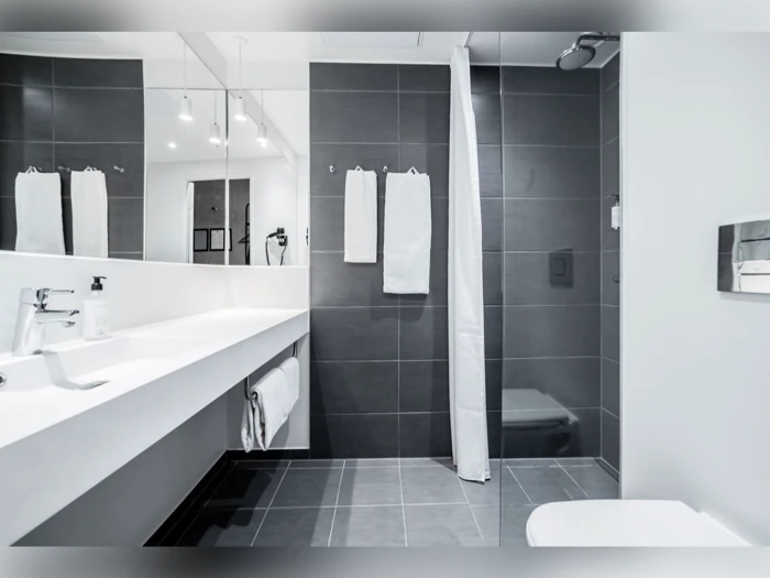 Moderne badeværelse med mørkegrå fliser, hvide bordplader, dobbelte håndvaske, et vægspejl, hvide håndklæder og en brusekabine med forhæng.
