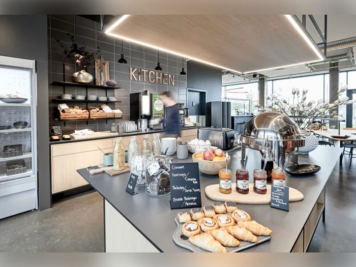 Moderne køkkencafé med kager, syltetøjsglas, frugt og morgenmadsprodukter på en disk. En person bevæger sig i nærheden af en kaffemaskine. Hylder, tallerkener og brød er synlige i baggrunden.