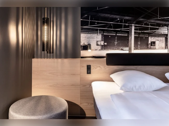 Moderne hotelværelse med en seng, hvidt sengetøj, en rund grå skammel, træpaneler, en hængelampe og et sort-hvidt industrielt foto på væggen.