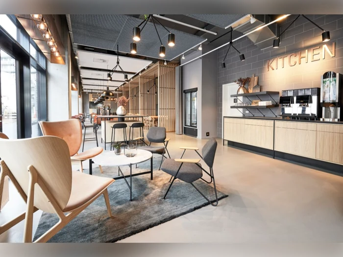 Moderne åbent kontorkøkken med stilfulde lænestole, kaffemaskine, borde og et stort KITCHEN-skilt på væggen; naturligt lys strømmer ind gennem store vinduer.