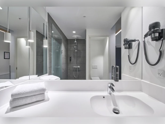 Moderne hotelbadeværelse med hvid håndvask, foldede håndklæder, væghængt hårtørrer, stort spejl og en glasindkapslet bruser med grå fliser i baggrunden.