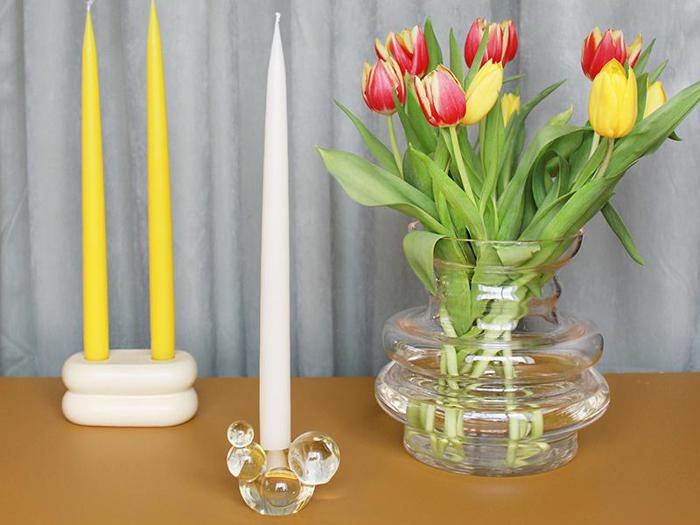 En glasvase med røde og gule tulipaner, et hvidt lys i en dekorativ glasholder og to gule lys i en moderne holder på et bord.