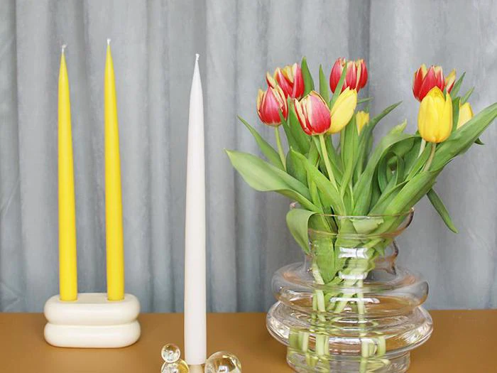 To gule lys i en keramikholder, et hvidt lys i en glasholder og en glasvase med røde og gule tulipaner på et bord foran grå gardiner.