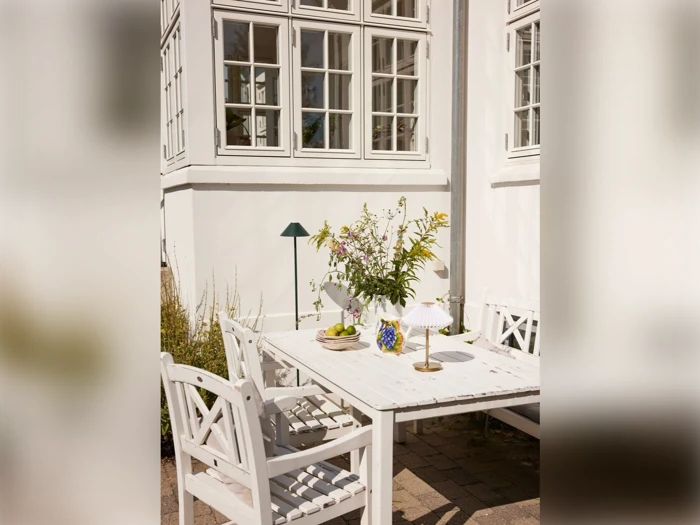 Et hvidt udendørs bord med stole er dækket med blomster, to lamper, en skål med frugt og glas, placeret foran en hvid bygning med store vinduer.