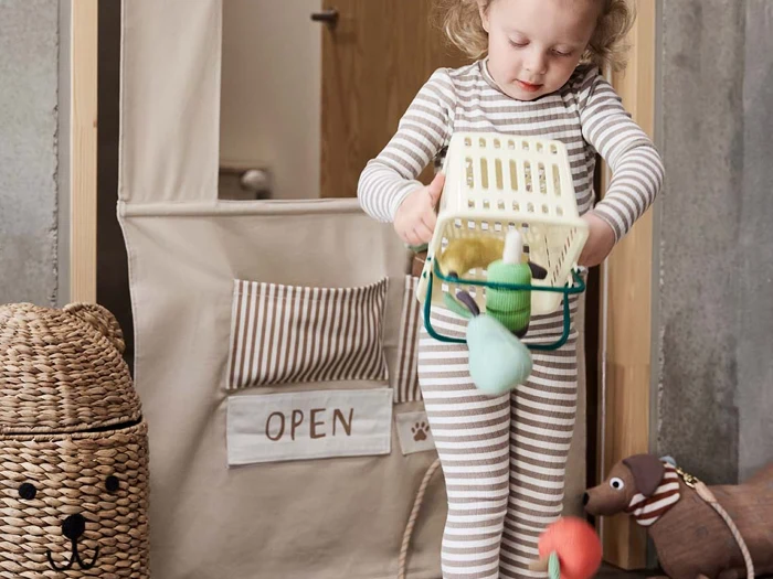 Et lille barn i stribet pyjamas holder en legetøjsindkøbskurv fyldt med dagligvarer, mens det står foran en legebutik med et ÅBEN-skilt.