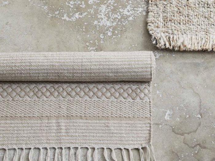To beige vævede tæpper på et slidt betongulv; det ene tæppe er rullet sammen og viser frynsede kanter og diamantmønstre, det andet er delvist synligt i hjørnet.