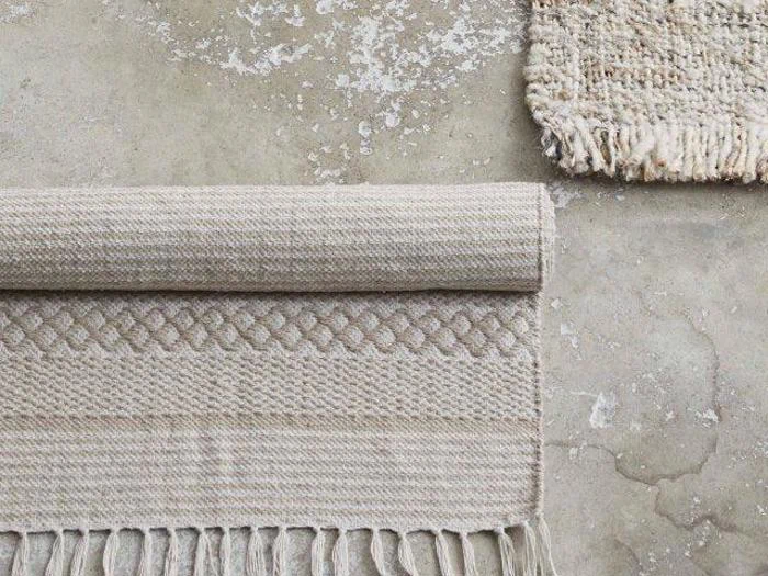 To beige vævede tæpper på et slidt betongulv; det ene tæppe er rullet sammen og viser frynsede kanter og diamantmønstre, det andet er delvist synligt i hjørnet.