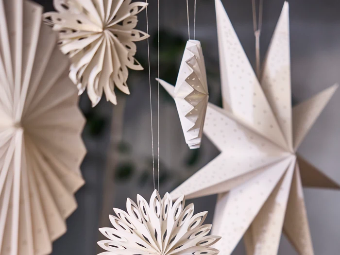 Papirdekorationer i forskellige geometriske former, bl.a. stjerner og rosetter, hænger i snore mod en sløret baggrund. Dekorationerne er hvide med fine udskæringsmønstre.