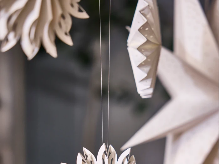Nærbillede af hvid papirpynt, der hænger i tynde snore, herunder stjerne- og blomsterformer, mod en sløret baggrund.