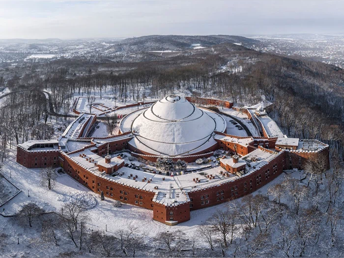 Luftfoto af en stor cirkulær fæstning med røde murstensvægge og et kuppeltag, omgivet af snedækkede træer og landskab om vinteren.
