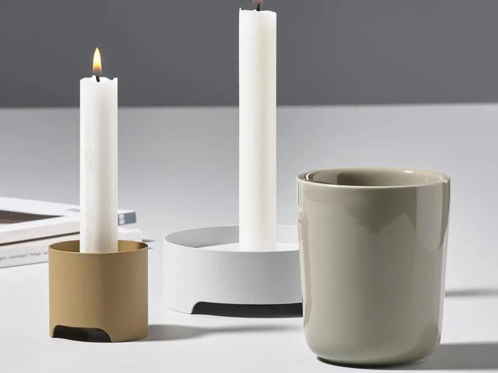 Tre minimalistiske lysestager, to med hvide stearinlys (et tændt og et slukket) og en grå kop på en hvid flade med en bog i baggrunden.