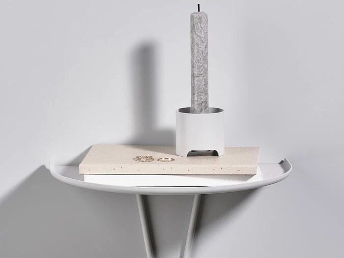 Et rundt hvidt bord med en beige bog, et par øreringe og et højt gråt stearinlys i en hvid holder på toppen mod en lysegrå baggrund.