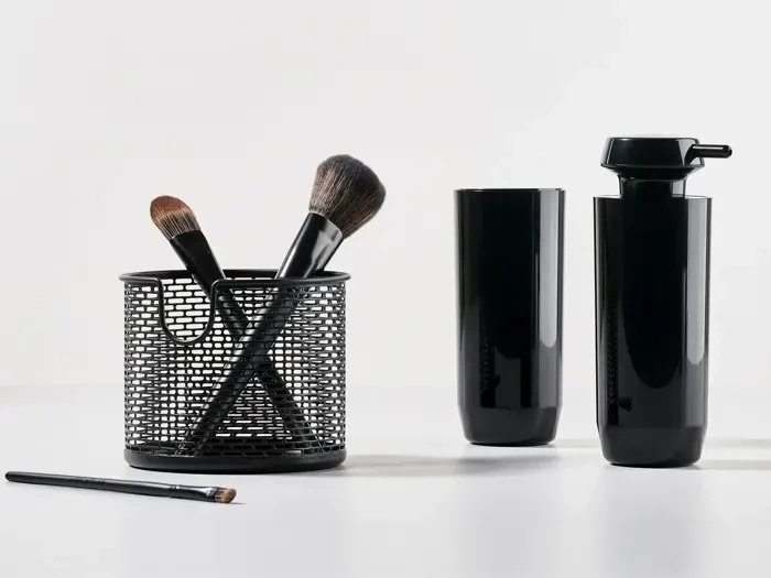 Sorte makeupbørster i en netholder, en makeupbørste, der ligger på bordet, og to slanke, sorte, cylindriske beholdere på en hvid overflade med almindelig baggrund.