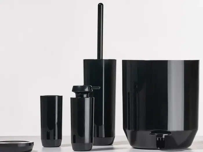 Et sæt skinnende sort badeværelsestilbehør, herunder en toiletbørsteholder, en sæbedispenser, en tumbler og en affaldsspand, arrangeret på en lys overflade mod en almindelig baggrund.