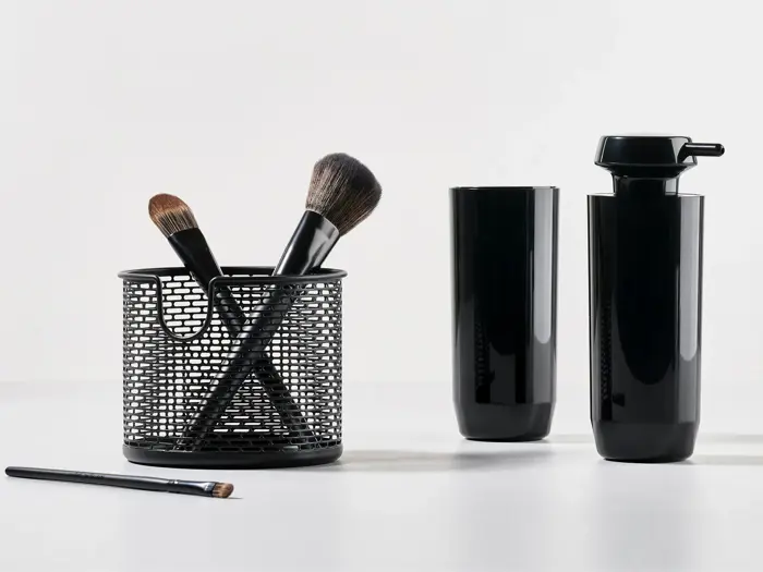 Sorte makeupbørster i en netholder, en makeupbørste, der ligger på bordet, og to slanke, sorte, cylindriske beholdere på en hvid overflade med almindelig baggrund.
