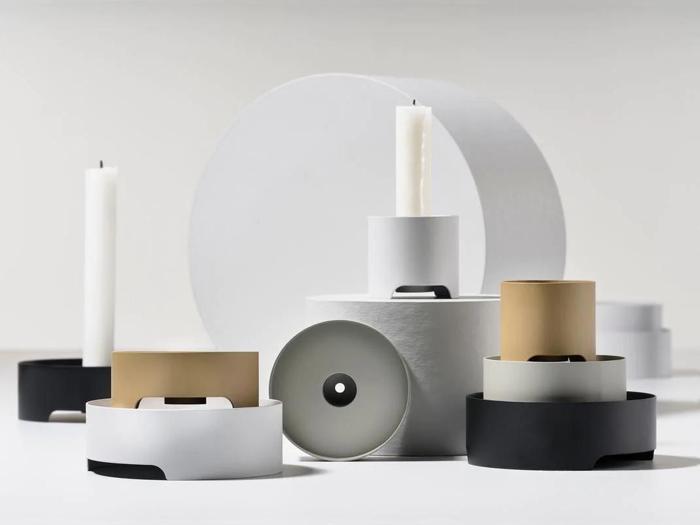 Minimalistiske lysestager og beholdere i hvide, sorte og gyldne toner er arrangeret på en hvid flade med stearinlys og geometriske former, der skaber en moderne, stilfuld komposition.