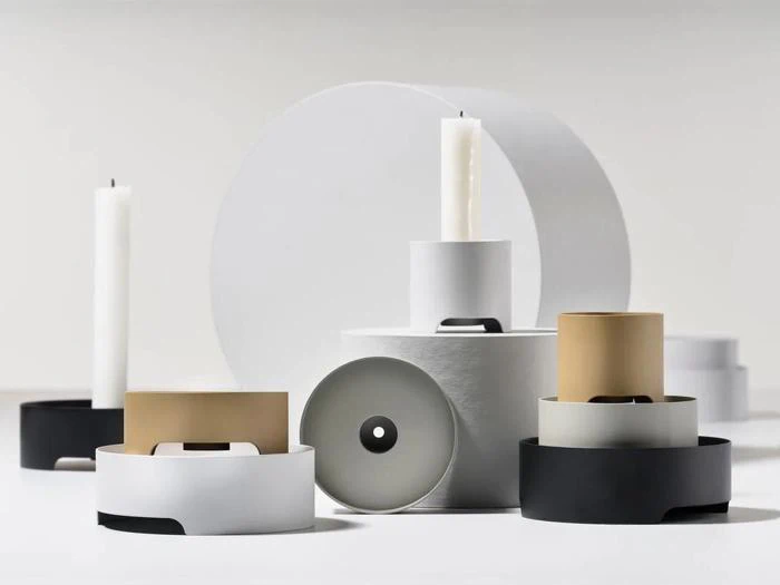 Minimalistiske lysestager og beholdere i hvide, sorte og gyldne toner er arrangeret på en hvid flade med stearinlys og geometriske former, der skaber en moderne, stilfuld komposition.