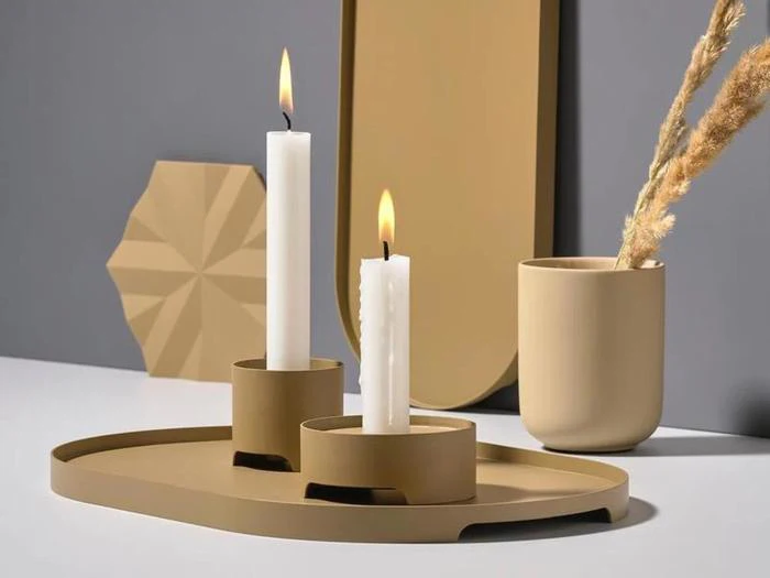 En moderne beige og brun indretning med to tændte hvide lys i holdere, en kop med tørret græs og geometriske former på en glat overflade mod en grå baggrund.