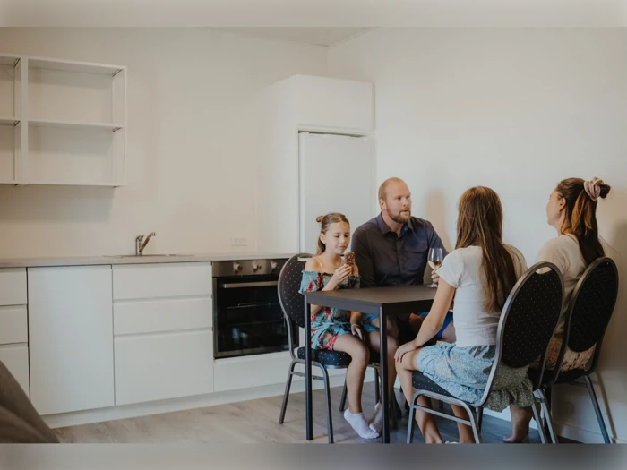 En familie på fire sidder omkring et lille bord i et moderne køkken og snakker og smiler. Rummet har hvide skabe og minimal indretning.