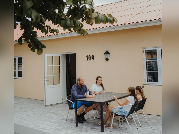 Fire personer sidder omkring et bord uden for et beige hus med nummeret 109 og taler og smiler. Døren er åben, og der er et træ delvist synligt til venstre.