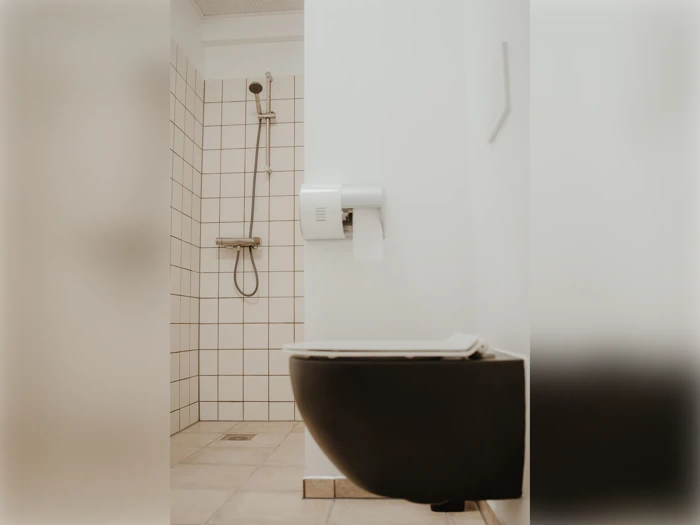 Minimalistisk badeværelse med et sort væghængt toilet, toiletpapirholder og en brusekabine med hvide, firkantede fliser og et håndholdt brusehoved.