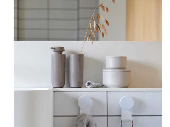 Minimalistisk badeværelsesbord med to taupefarvede beholdere, et rør, to stablede runde beholdere, tørret græs i en høj kop og to kroge med håndklæder hængende under.