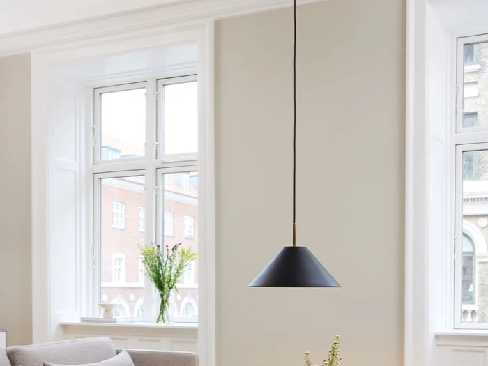 En moderne sort pendellampe hænger foran store vinduer i en lys, minimal stue med lyse vægge og en grå sofa.