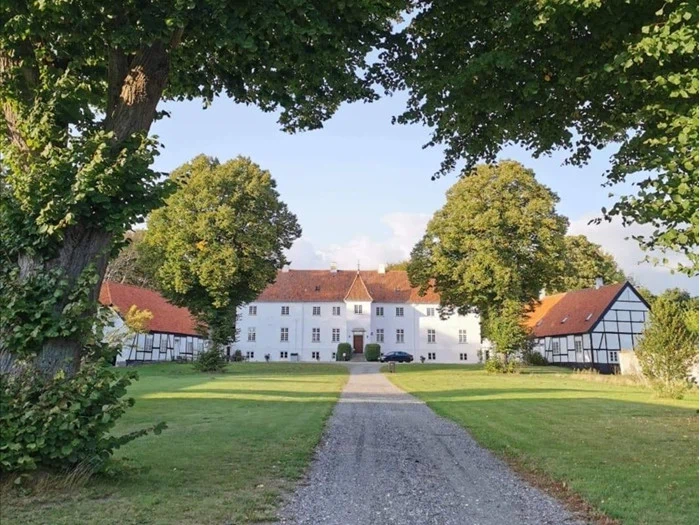 En bred grussti fører til en stor hvid herregård med røde tage, omgivet af grønne plæner og høje træer under en klar blå himmel.