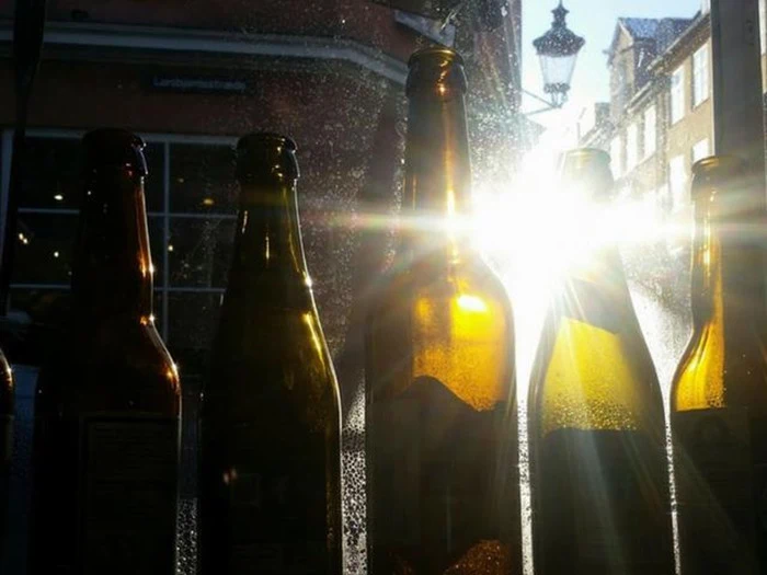 Sollyset strømmer gennem flere tomme glasflasker i en vindueskarm og skaber lyse refleksioner og silhuetter mod en urban bygningsbaggrund.